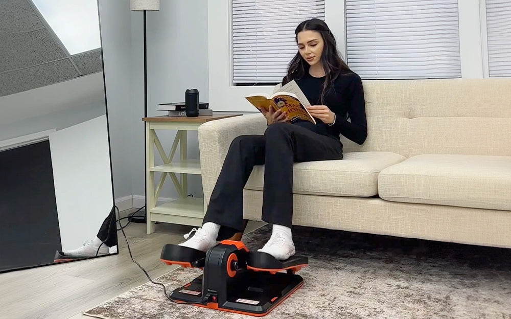 the female is using the mini elliptical machine while reading the book